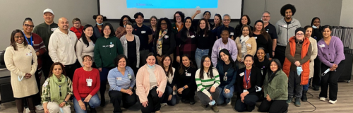 A large group of people pose together in a meeting room during a professional development event, with some standing and others kneeling in front. A presentation screen is visible behind them, and many participants wear name badges.