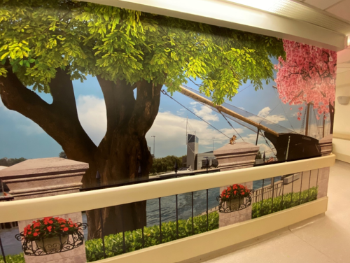 A long hallway wall features a large, vivid art installation showing a leafy tree, flowering branches, stone railings with planters, and a waterfront scene with city buildings in the background, creating a bright, outdoor‑inspired atmosphere indoors.