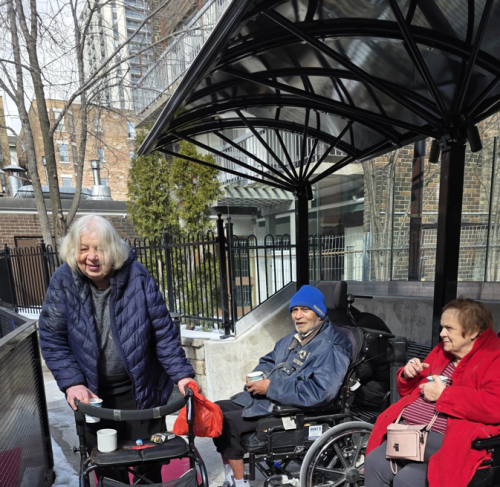 Three residents use the new outdoor, open-sided shelter at Fudger House. Two residents sit in wheelchairs, while one stands and pushes their walker.