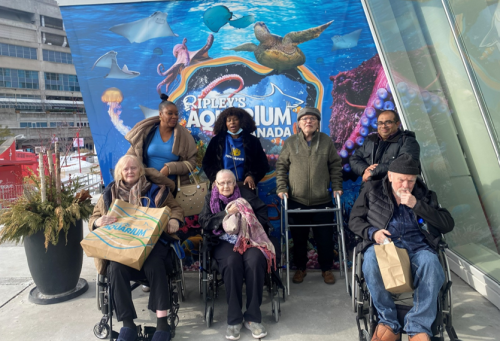 Three staff and four residents from True Davidson Acres stand together outside Ripley’s Aquarium, posing in front of a bright blue wall decorated with fish and underwater imagery.