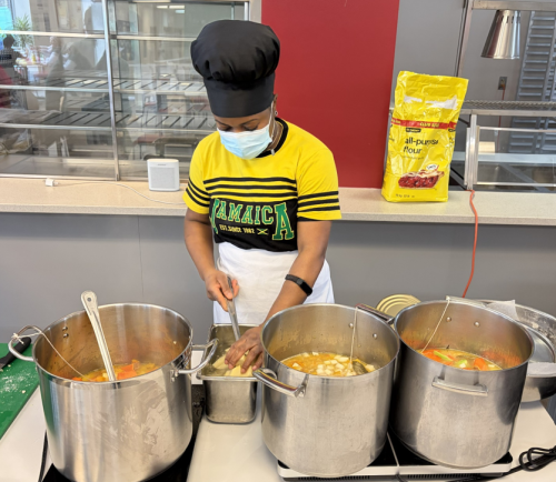 A cook wearing a yellow shirt that says “Jamaica” and a black chef hat prepares food at a counter, stirring ingredients in large metal pots on portable burners during a cooking demonstration.