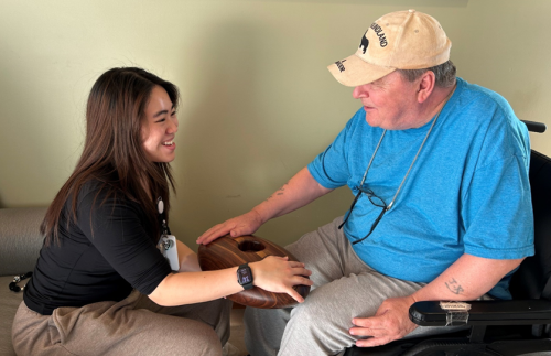 A resident and a staff member sit together with their hands on a wooden instrument, which uses sound and touch to deepen connection. The resident sits in a wheelchair and smiles at the staff member, who is sitting across from him.