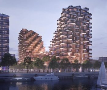 Photograph of modern waterfront residential buildings with unique, staggered balcony designs illuminated at dusk. Boats and trees line the water in foreground.