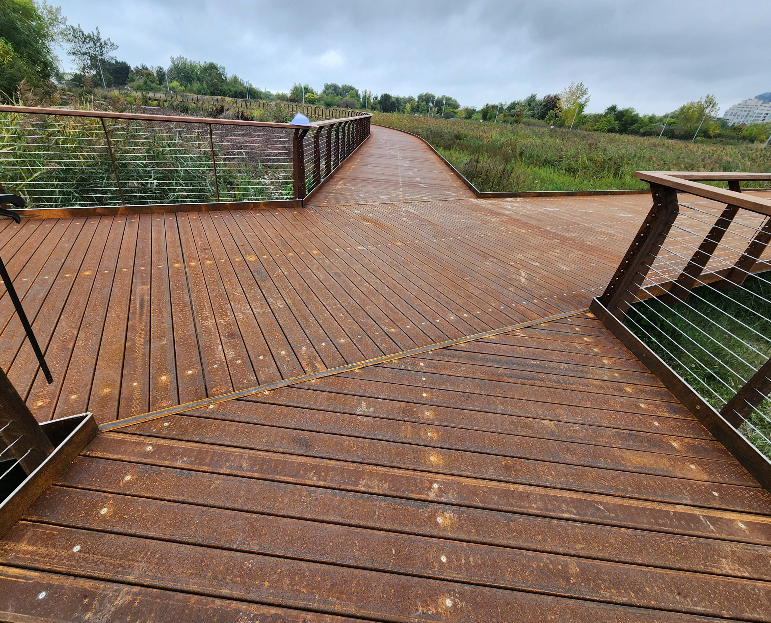 Wide view of a metal boardwalk intersection with wooden handrails and horizontal cabling on an overcast day. Dense vegetation surrounds the boardwalk, and trees are visible along the horizon.
