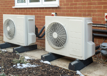 Heat pumps outside of a red-brick home on concrete with stones and snow nearby.