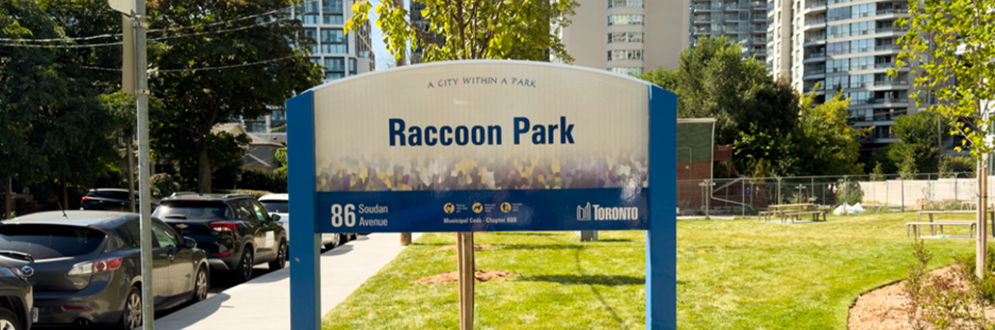 Sign for Raccoon Park in Toronto with grassy area, trees, and apartment buildings in the background.