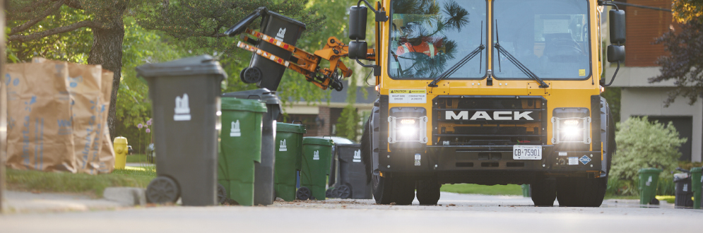 Garbage Bins, Green Bins and yard waste set out by the curb on a residential street. A collection truck is lifting a Garbage Bin with its mechanical arm.