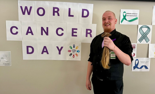 Josh stands holding his newly cut ponytail in front of a wall that says “World Cancer Day,” marking a Cancer Awareness Month event where he donated his hair in support of children affected by cancer.