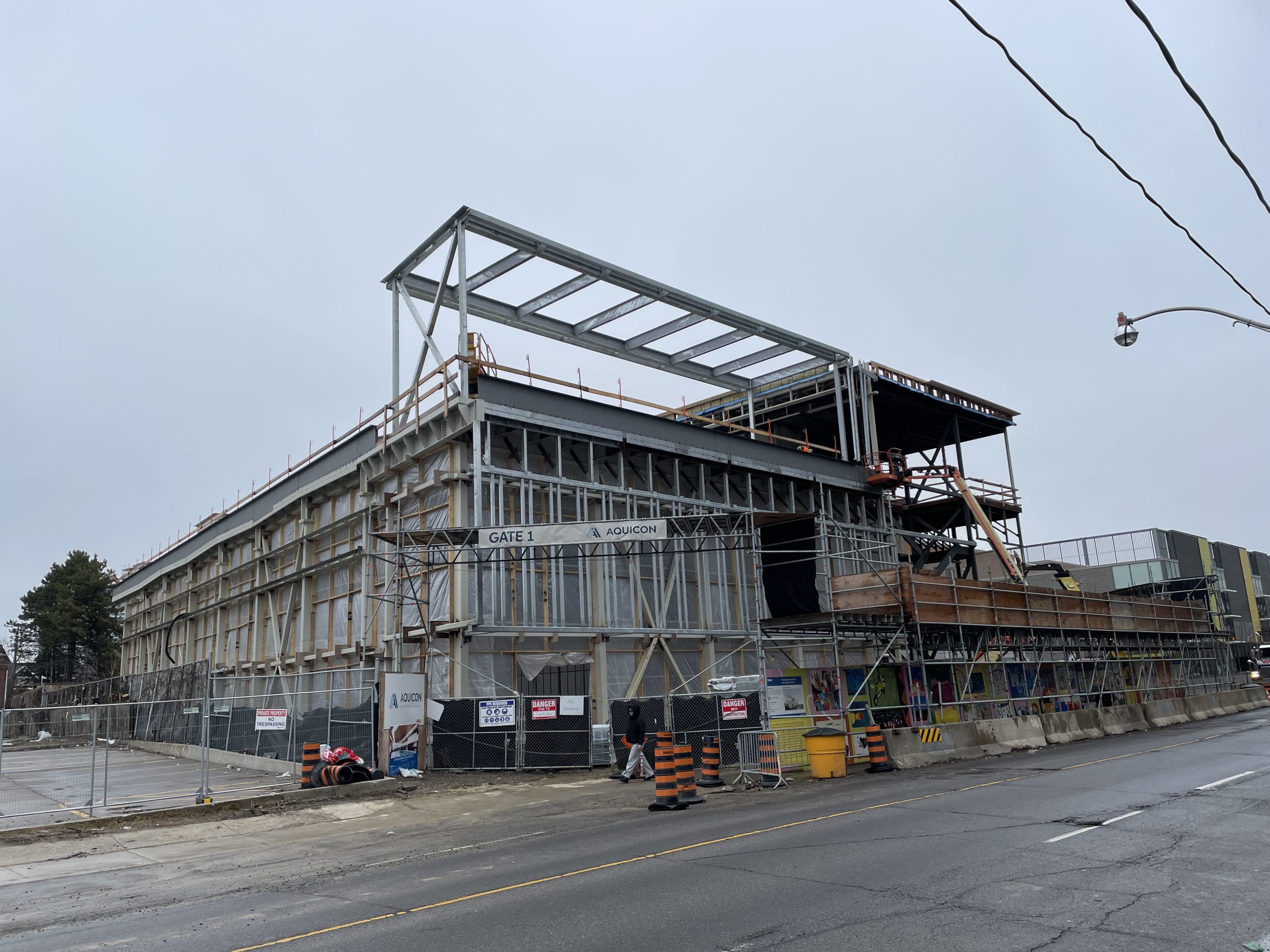 A view of a construction site for a multi-storey building on a cloudy day. Steel framing and scaffolding make up the building’s structure, with exterior walls partially complete. Construction fencing and hoarding surround the site. 