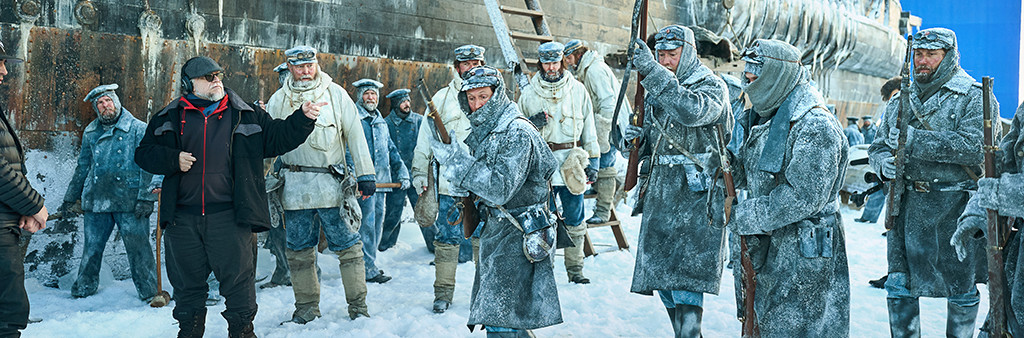 Actors on set of an old ship in winter time dressed in costumes holding muskets with a director point at something.