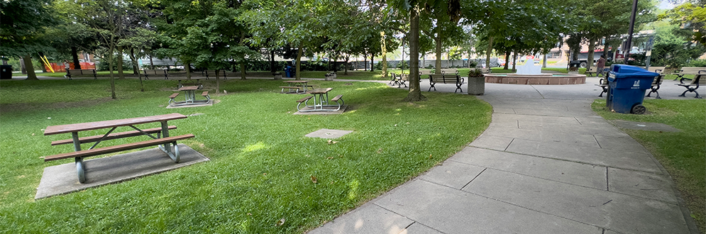 A park area with several picnic tables placed on a grassy lawn surrounded by mature trees. A paved pathway curves along the right side of the image, with a blue recycling bin and a stroller visible near the path. The background includes more trees, open space, and a glimpse of a playground area.