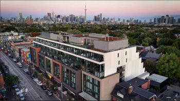 Aerial photograph of a modern multi-story building with large windows and balconies, situated in an urban neighborhood with tree-lined streets. City skyline with tall skyscrapers and a prominent tower is visible in the background during sunset, highlighting a blend of residential and commercial architecture.