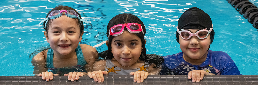3 kids in swim goggles in a pool hanging onto the edge and smiling for the camera.