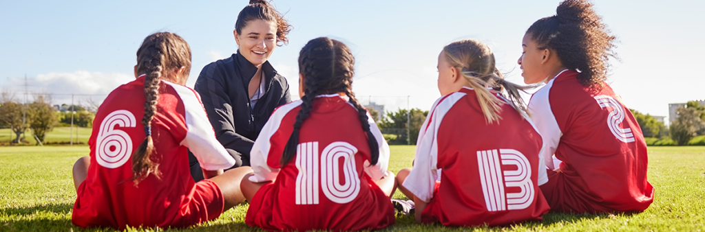 Four young girls wearing soccer jerseys sit in a circle with their female coach.