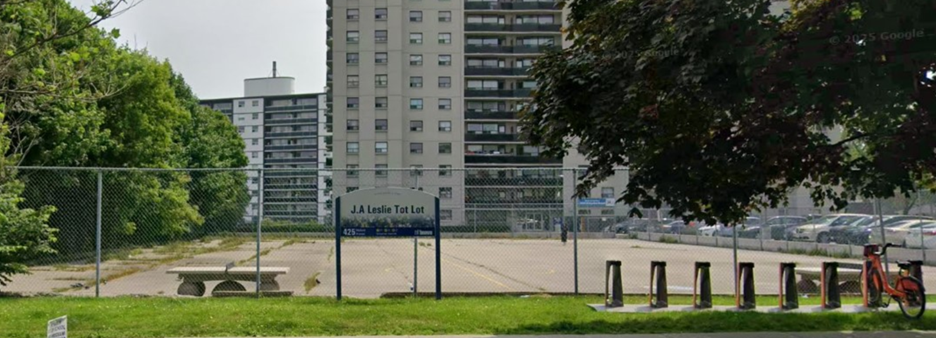 A view of J.A. Leslie Tot Lot from Midland Avenue, facing east showing a large concrete pad with a tall fence around it and a tall apartment building in the background.
