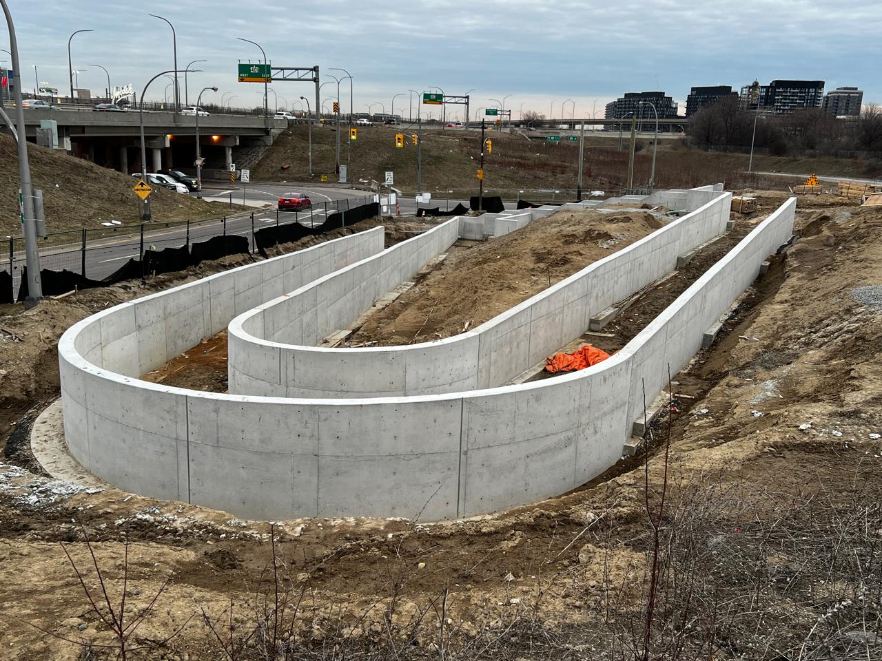 View of the construction site highlighting progress on the ramp and stairs. The concrete foundation and structural walls are fully cast. Between the two parallel retaining walls are soil and construction materials. Vehicles and a roadway are in the background.