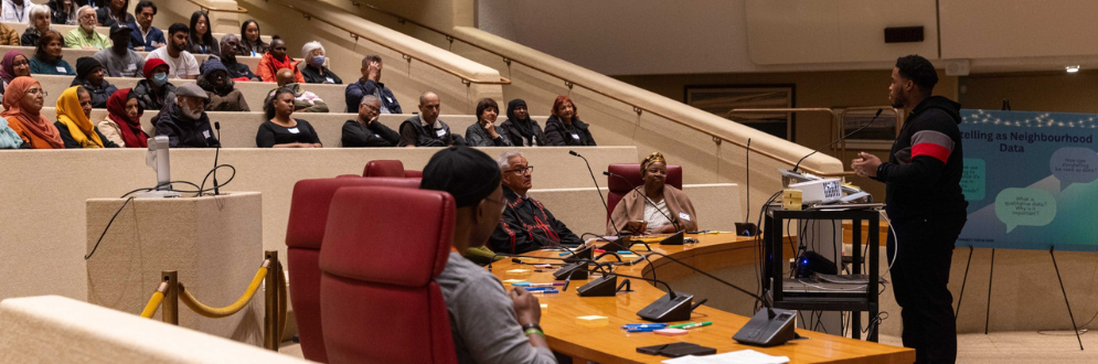 A community leader stands at the front of a large council‑style chamber, speaking to an audience seated in tiered rows. The speaker stands beside presentation equipment and addresses attendees who listen attentively.