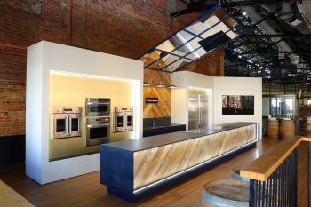 Indoor image of St. Lawrence Market Kitchen, showing a long island infront of a rentable kitchen space with multiple ovens on the left side a sink in the middle and a large kitchen and freezer on the right. A television is also seen at the far right side.