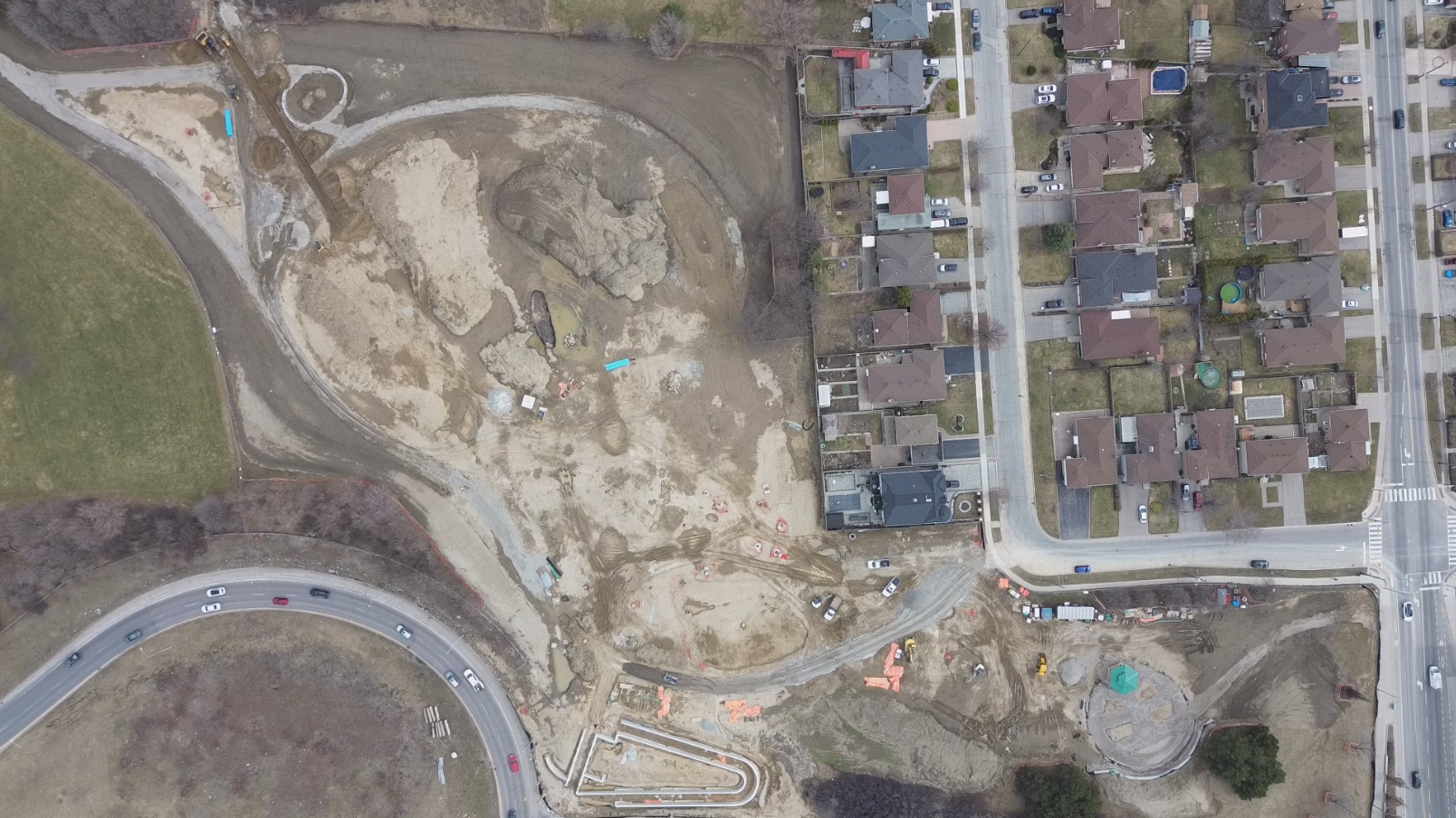 Aerial view of the construction site highlighting overall progress. Construction materials and vehicles are scattered throughout the large construction site. The surrounding area includes residential houses, roads and grass fields.