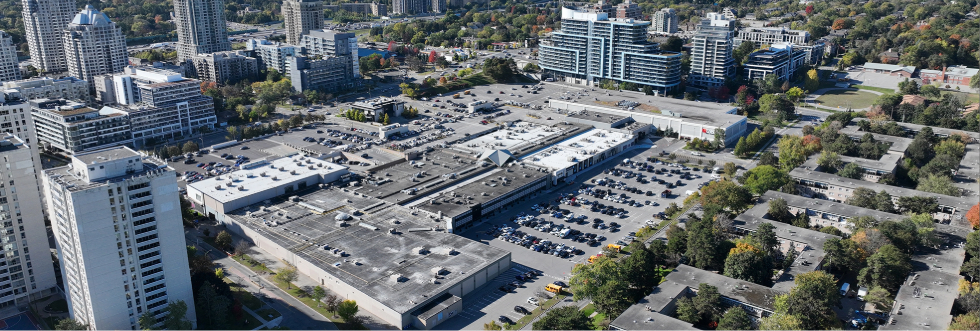 A drone level overview photo looking south west at the future park site. The photo is taken from above Hawksbury Park near the intersection of Hawksbury Drive and Bayview Mews Lane. The photo includes the Bayview Mews Apartments the north of the site and the Bayview Village Shopping Centre to the south. Residential towers in the distance surround the shopping centre and parking areas.