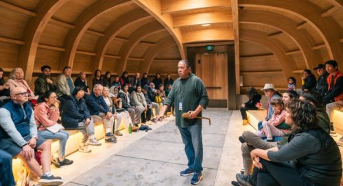 Seated audience on left and right of a central aisle where a standing person speaks to the audience, inside an arched wooden room