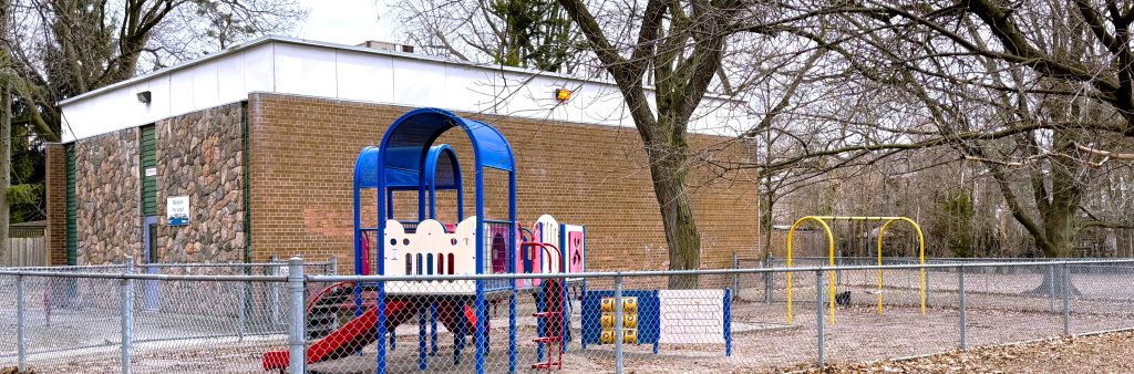 Photo of the current playground at Cedarvale Park located within a fenced area. It features one main play structure with two slides, climbing structures and play panels, and one swing set.