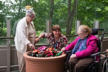 Two older adults and one younger participant gathered outdoors around a large planter, tending to colourful flowers, with one participant seated in a wheelchair and trees and greenery in the background.
