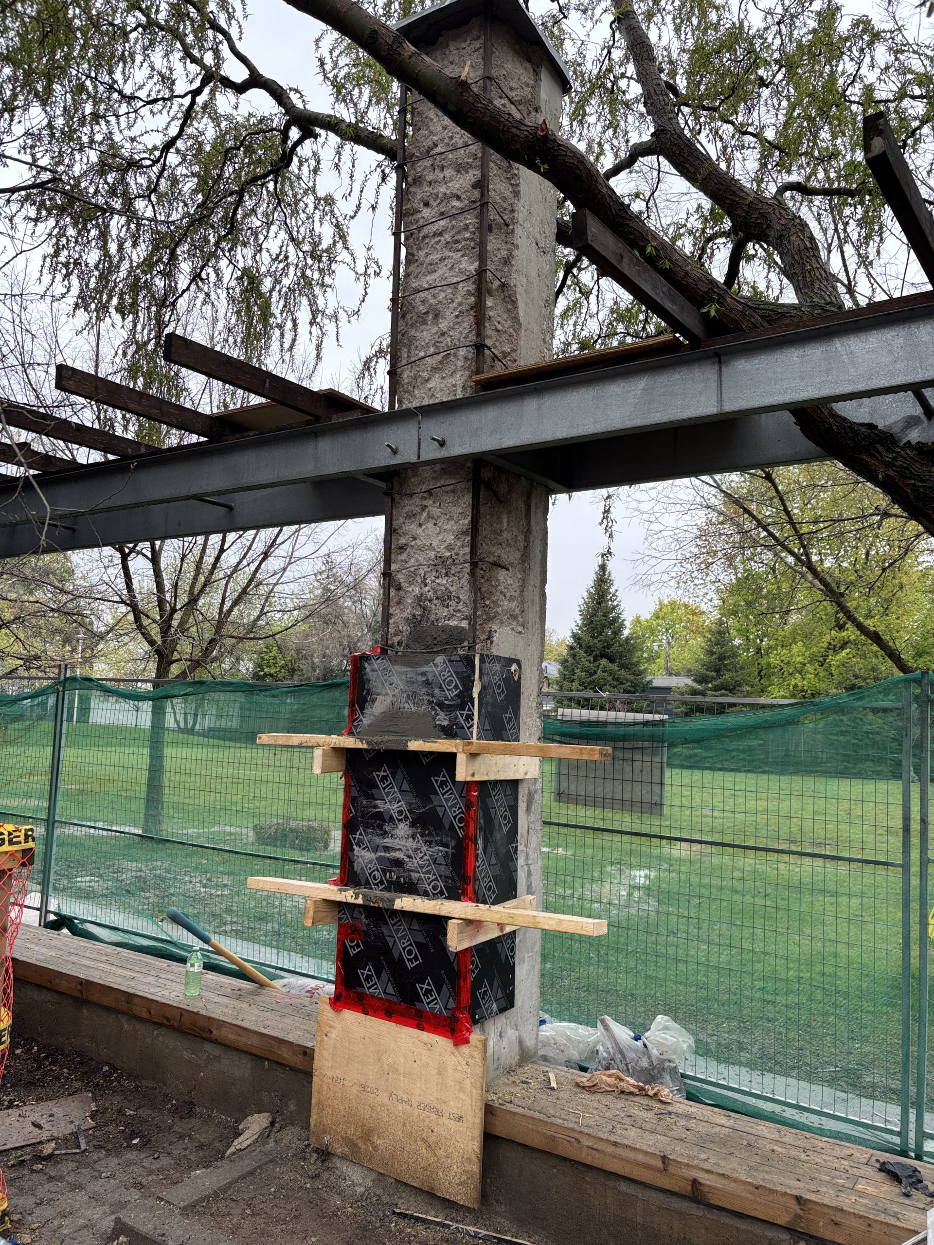 A close-up of a concrete column under construction at Wychwood Barns Park. Black panels are secured onto the lower half of the column by wooden slats and tape. Horizontal steel beams span above. The site is enclosed by a green mesh fence, with construction materials visible in the foreground.