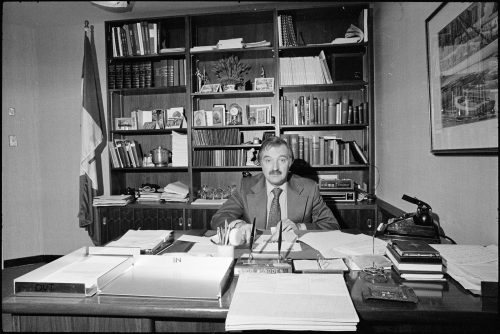 Image depicts man with a moustache sitting behind office desk.