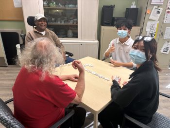 Four people sitting around a table in a shared room, playing dominoes together, with two participants wearing face masks and one using a wheelchair, and cabinets and equipment visible in the background.