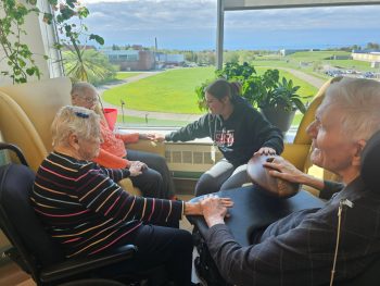 Small group of residents seated by a large window participating in a guided sensory activity, with a staff member supporting interaction using a handheld wooden device.