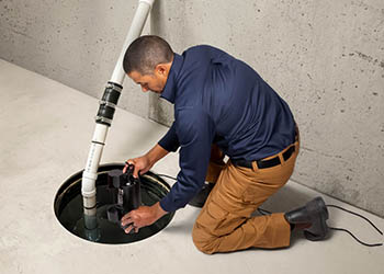 A person kneeling on a basement floor while adjusting a sump pump inside a circular sump pit.