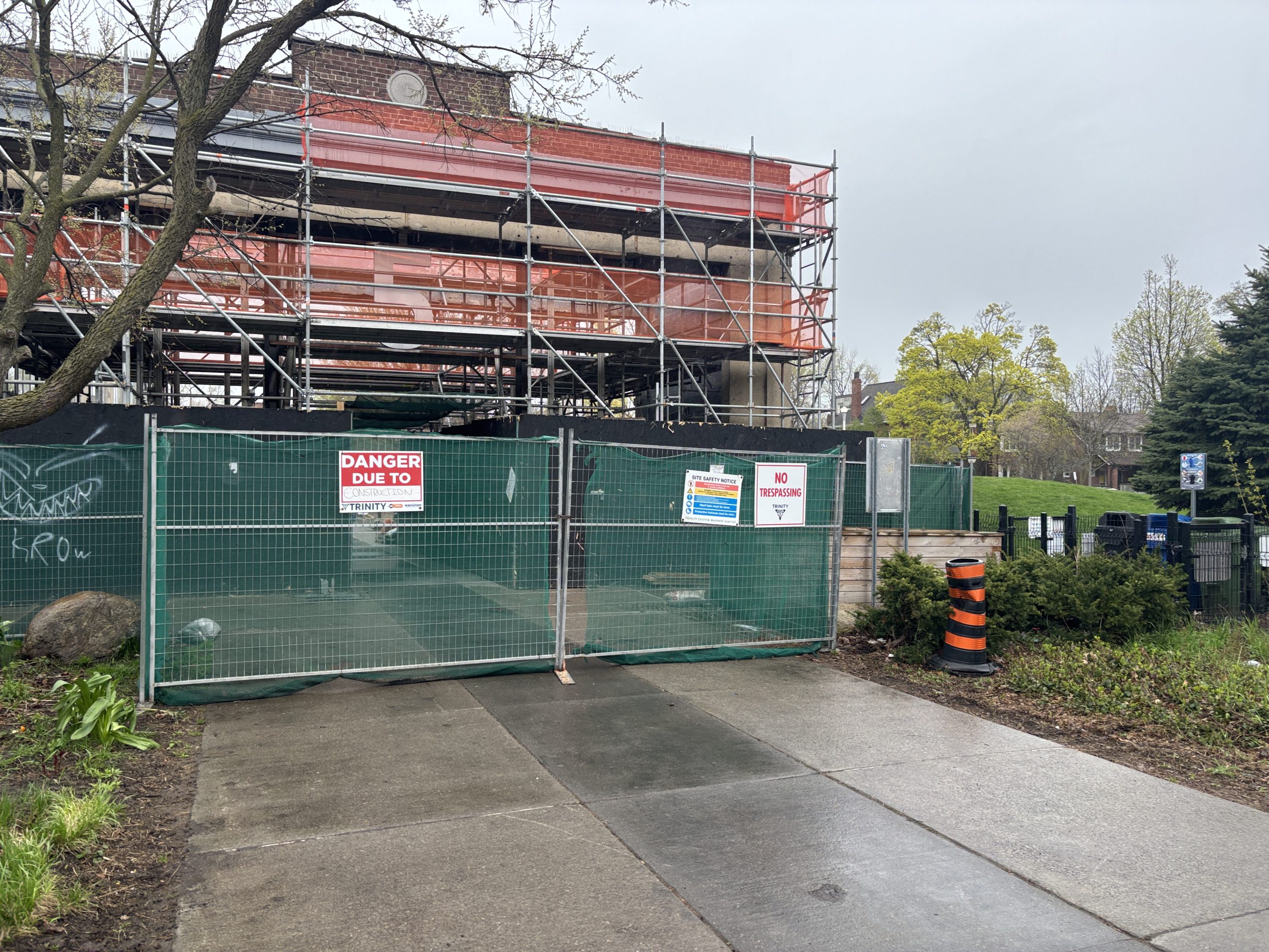 A construction site at Wychwood Barns Park showing Barn 5 surrounded by worker scaffolding. A green temporary fence with "Danger Due to Construction" and "No Trespassing" signs is installed along the perimeter. A paved pathway runs along the front of the fencing.