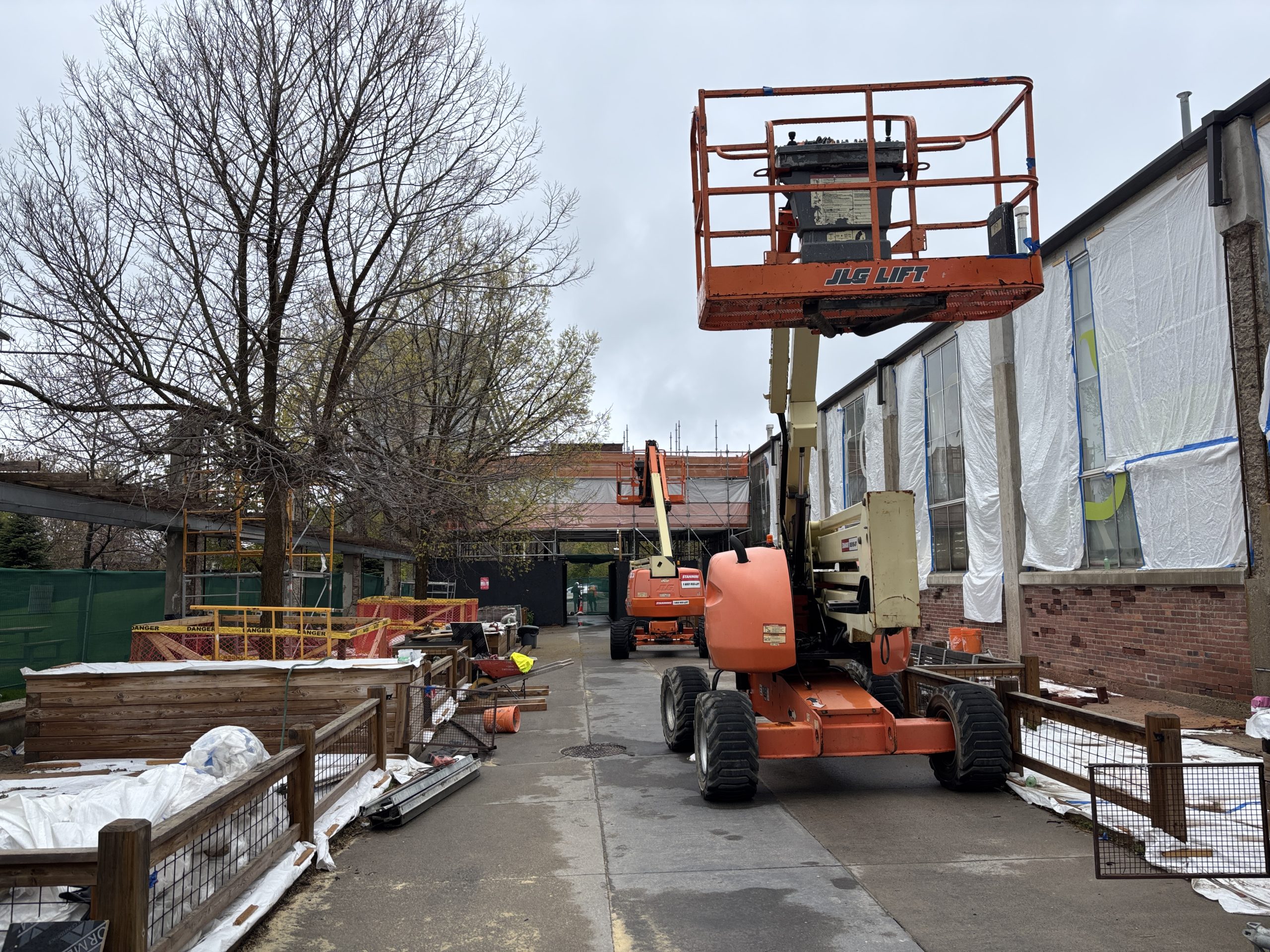 A construction site at Wychwood Barns Park with a construction lift parked on a paved path, beside a building under renovation. The building’s windows are covered in plastic sheets. Another lift is visible in the background near scaffolding. Construction materials are strewn along the path.