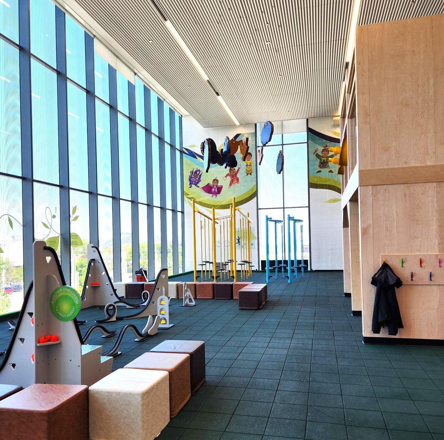View from the north-east corner showcasing the build-your-own coaster activity station, a corner of the house feature with jacket hooks, and suspended clouds and birds from the ceiling. In the background, stilts in two colours and a mural on the west wall are visible. The floor is finished in green rubber.