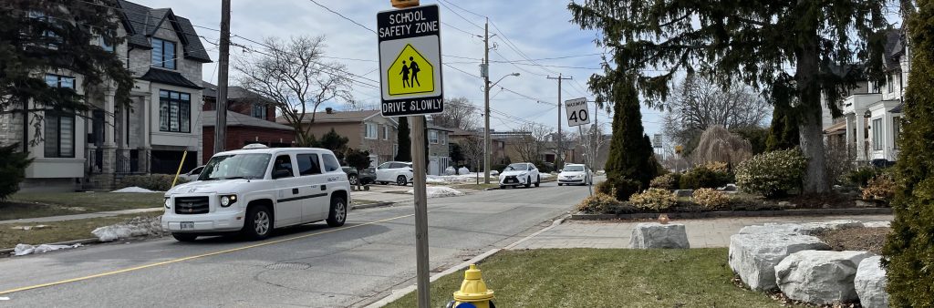 School safety zone sign with flashing beacon and “Drive Slowly” message on a residential street with passing cars and houses.