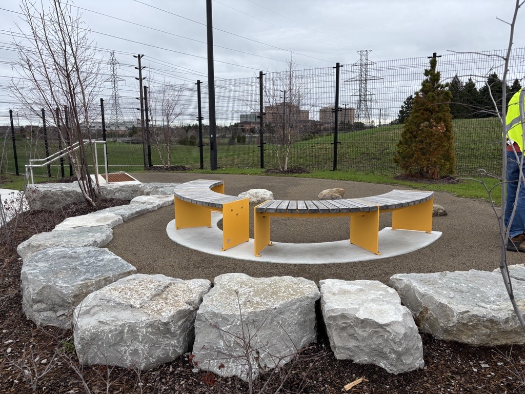 A circular seating area in a park featuring curved benches with yellow metal supports and wooden slats, set on a concrete base and surrounded by large natural boulders. A grassy hill, metal fencing, and hydro towers are visible in the background under an overcast sky.