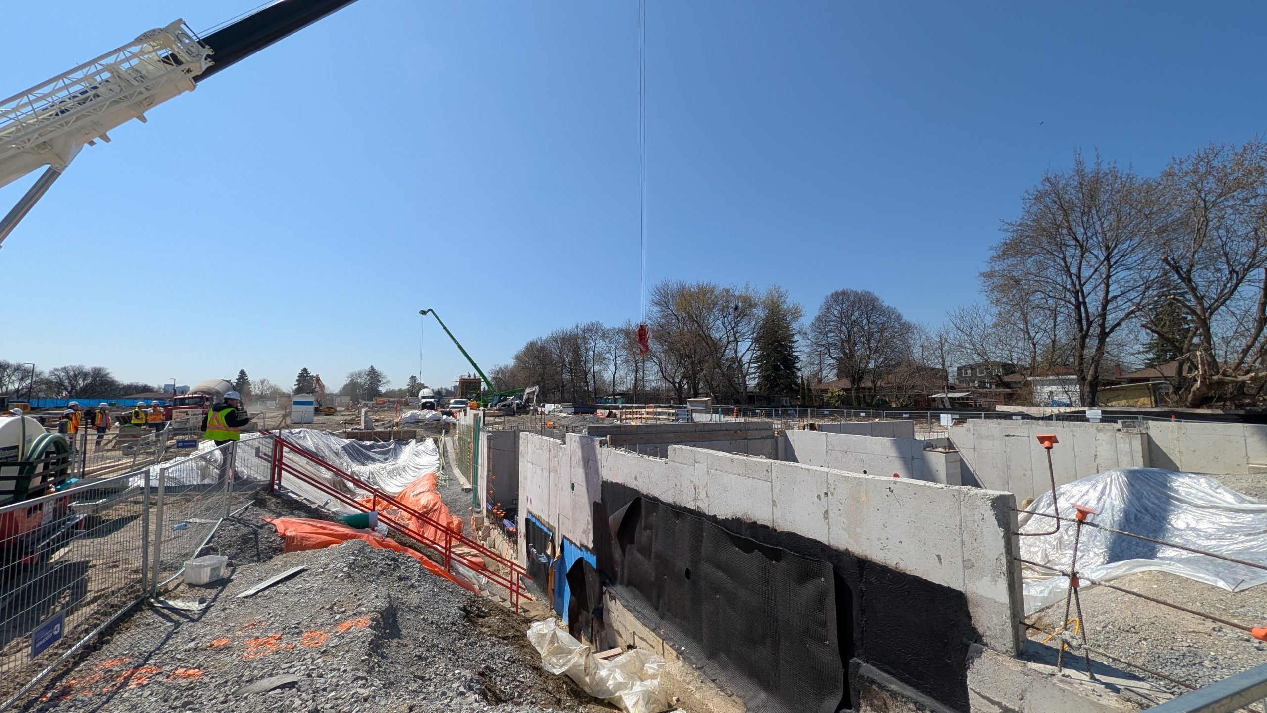 A section of an active construction site with concrete foundation walls, waterproofing and temporary barriers along a gravel edge. Workers in safety vests and hats stand nearby, surrounded by construction equipment, tarps and other materials. Trees and buildings are visible in the background under a clear blue sky.