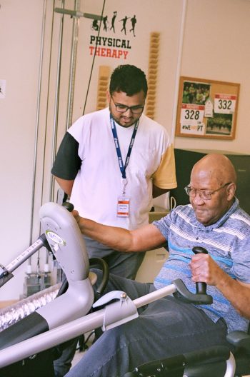 Older adult using a stationary exercise bike during a physical therapy session, while a staff member wearing an ID badge stands nearby providing support; “Physical Therapy” poster and bulletin board visible in the background.