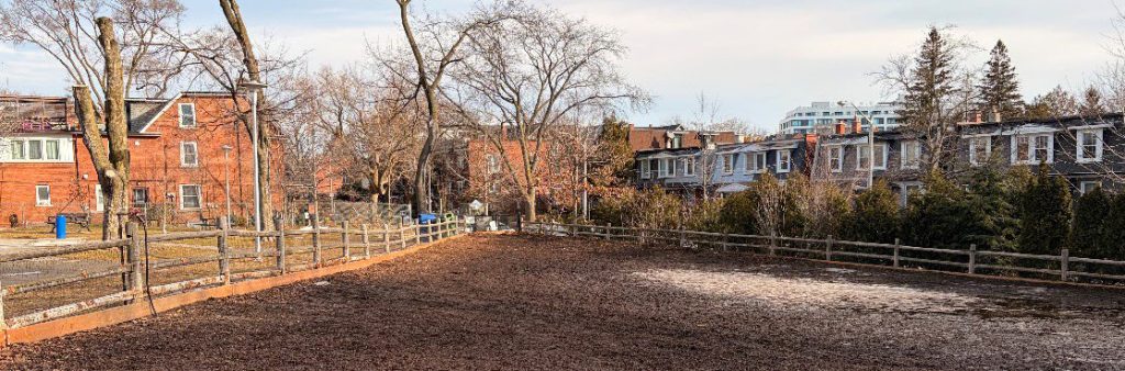 An open off-leash dog area in Baird Park, with a wood chip ground surface, wooden post-and-rail fencing, and bare trees, surrounded by brick rowhouses and low-rise residential buildings