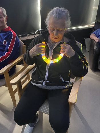 Older adult seated in a chair holding a glowing, multicoloured sensory tube, seated in a dimly lit room.