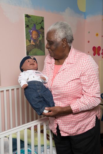 Older adult holding a lifelike baby doll beside a crib in a room decorated with child-themed wall art.