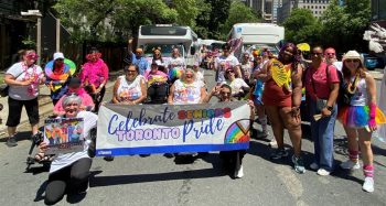 Group of older adults and volunteers participating in the Pride parade, holding a banner that reads “Celebrate Seniors Toronto Pride.” A city street with vehicles and buildings in the background.