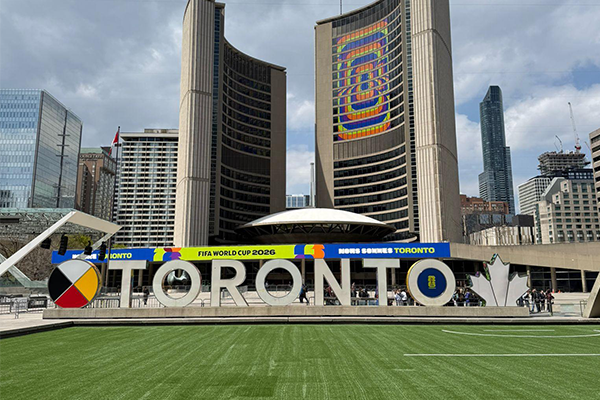 Nathan Phillips Square in front of Toronto City Hall with FIFA World Cup branding on the building and TORONTO Sign. A soccer pitch is featured in the front as well.