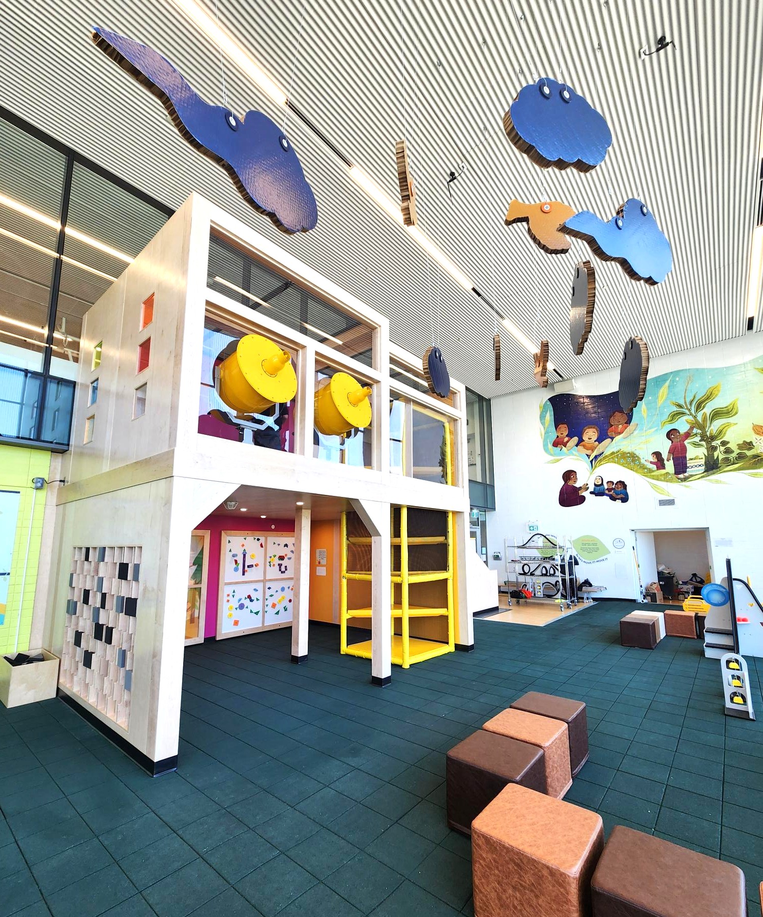 View from the south-west corner showcasing the house feature, suspended clouds and birds from the ceiling, a build-your-own coaster activity station, and a mural on the east wall. The floor is finished in green rubber. 