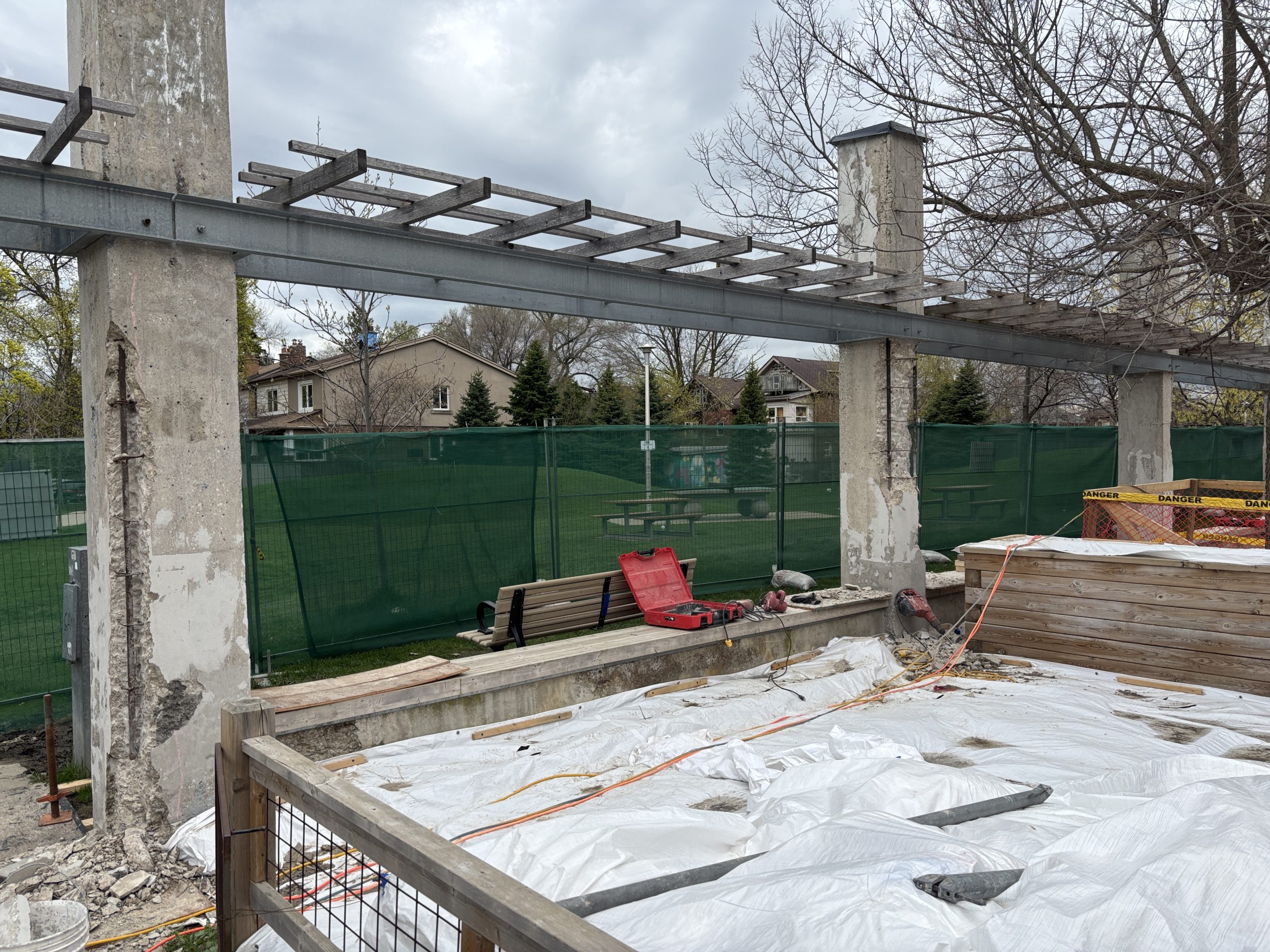 An active construction area inside the Wychwood Barns Park site. Concrete columns supporting a pergola structure show exposed masonry with paint removed. Construction tools, materials, and protective sheeting are visible on the ground. A green temporary fence surrounds the perimeter of the work zone.