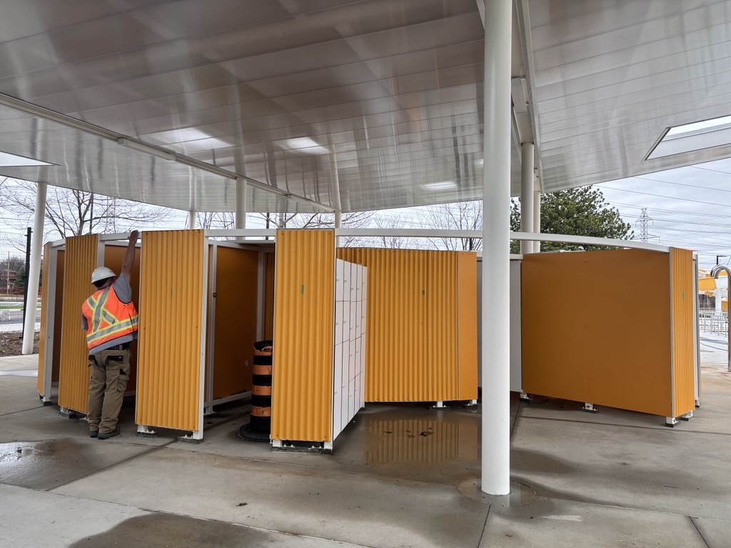 A construction worker in a high-visibility vest and hard hat inspects yellow corrugated changeroom or washroom stalls under a large modern canopy structure with a reflective metal ceiling. A construction pylon is visible inside one of the open stalls.