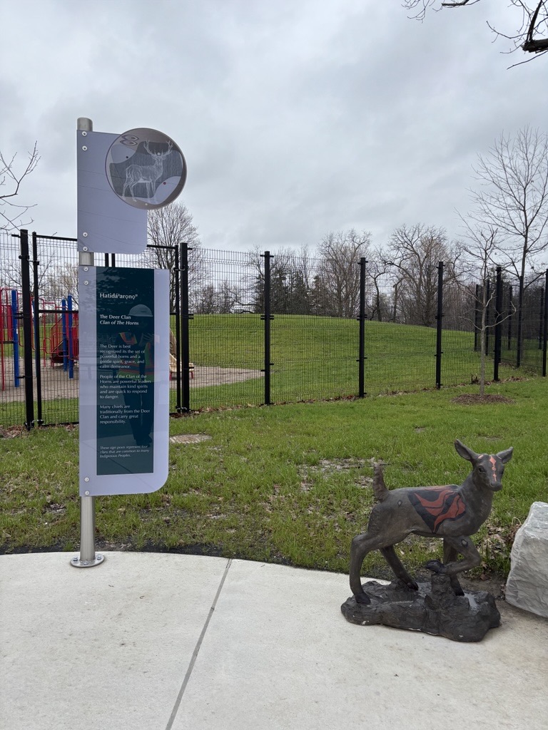An Indigenous placekeeping sign on a metal post featuring information about the Deer Clan, known as Hatida'aɔnɔ' or Clan of the Horns, with a circular deer motif at the top. A bronze deer sculpture with red painted markings stands beside the sign, with a grassy hill and park fencing in the background.