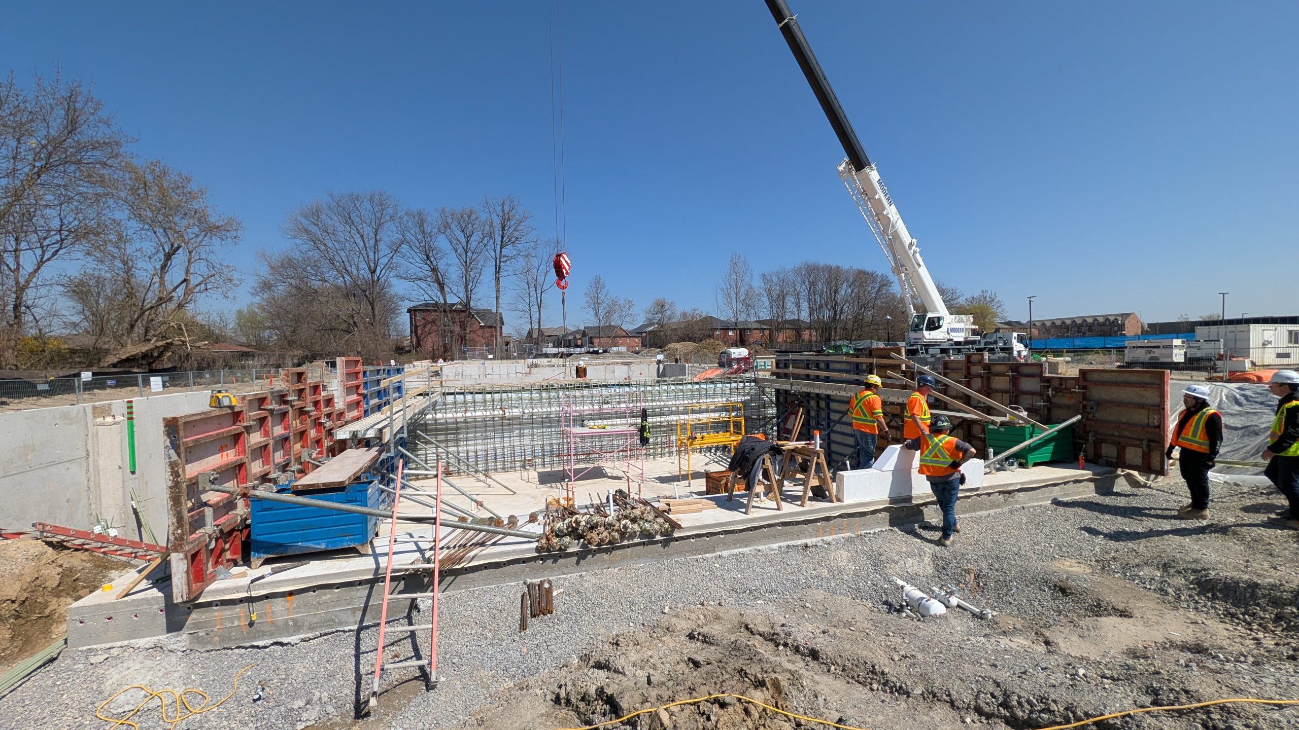 A wide view of an active construction site on a clear day. Workers wearing safety vests and hats stand along one edge of a deep excavated area with concrete foundation walls and formwork. The surroundings include scaffolding, construction materials and equipment. A mobile crane is visible in the background. 