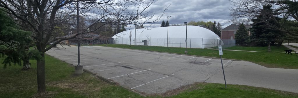 Empty parking lot in a park with a white inflatable dome structure enclosed by chain-link fencing in the background, surrounded by trees with spring foliage and a partly cloudy sky.